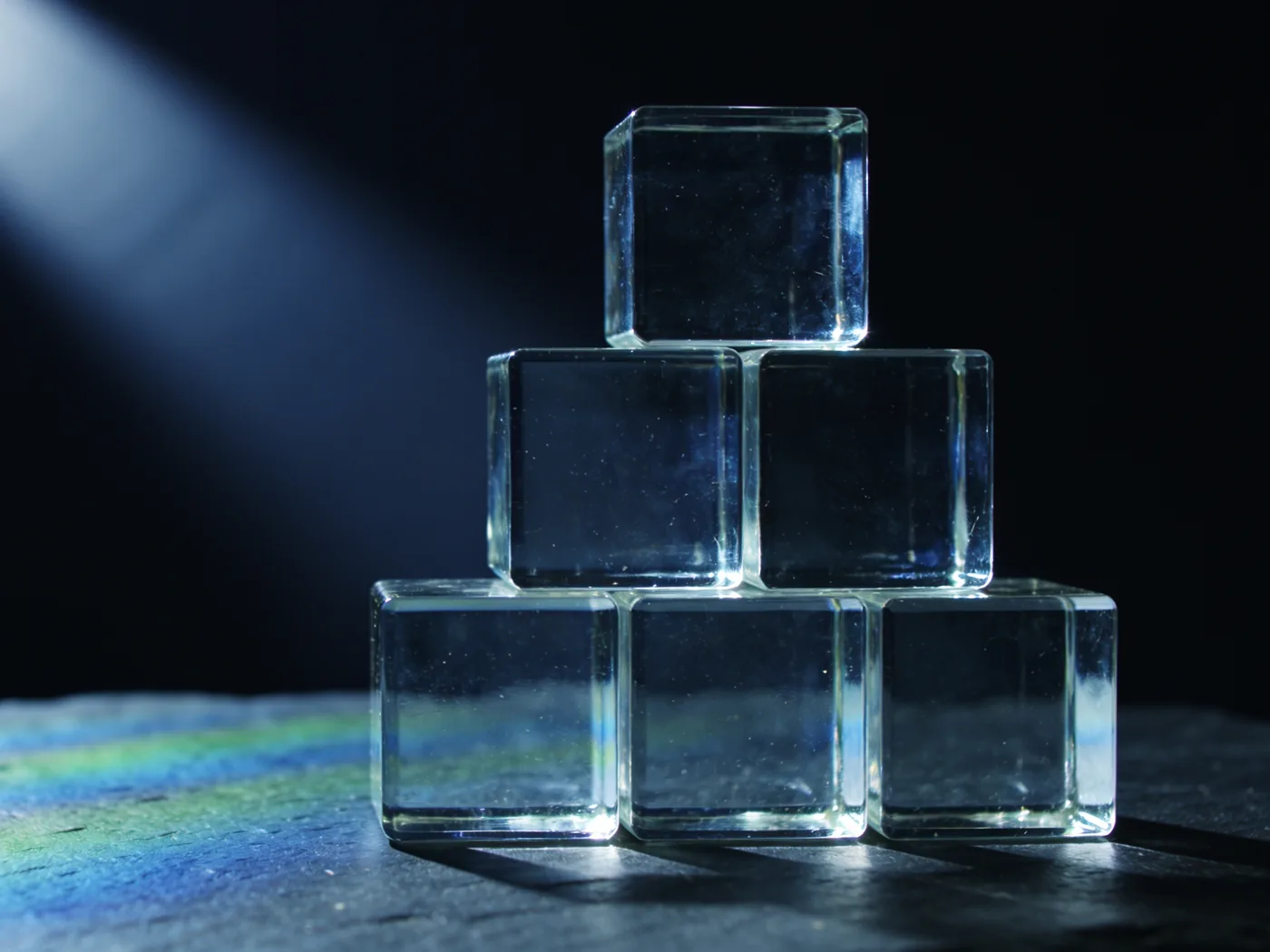 Sechs klare Glaswürfel, zu einer stabilen Pyramide gestapelt, auf dunklem Schiefer; ein Lichtstrahl zerlegt sich in Blau und Grün.